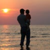 A silhouette of a father holding his son at the shoreline watching a sunset.