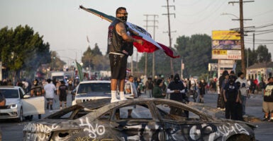 A masked protester stands atop a destroyed car waving a Mexican flag as agitators take over Paramount